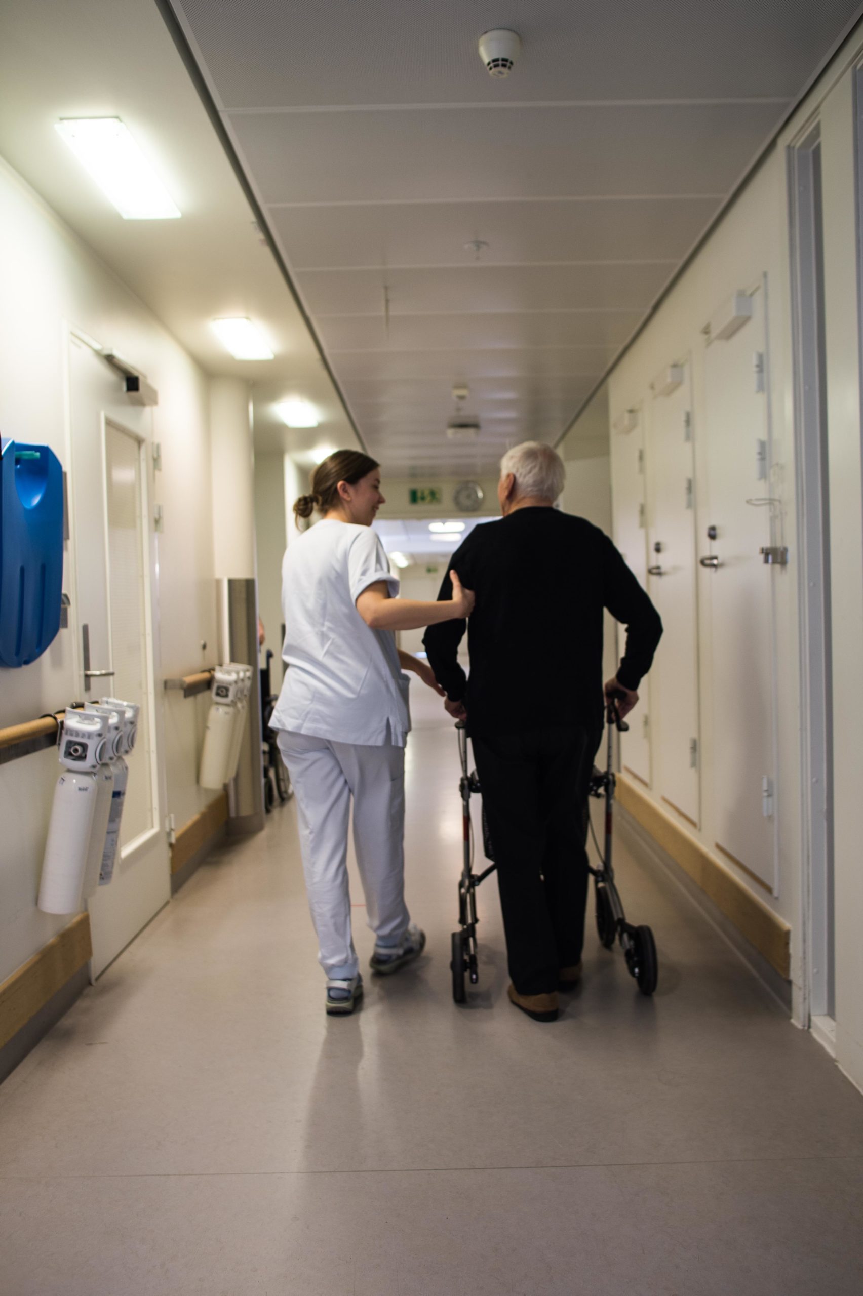 Sykepleier og eldre pasient med rullator går sammen i sykehuskorridor